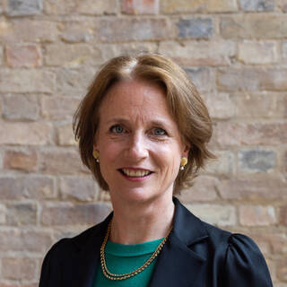 Colour photograph of the head and shoulders of a woman with blonde hair wearing black jacket. 